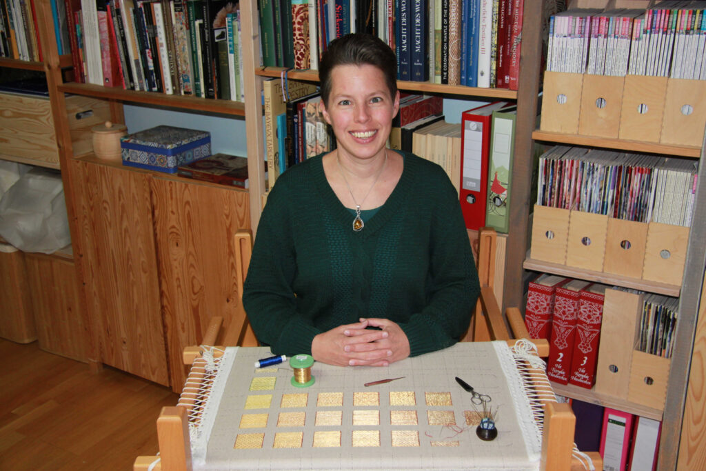 Acupictrix - Dr Jessica Grimm sitting behind her slate frame dressed with medieval goldwork embroidery.